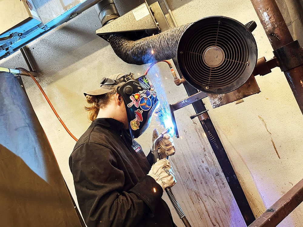 welding student, Mason Fisher, welds metal in a workshop setting.